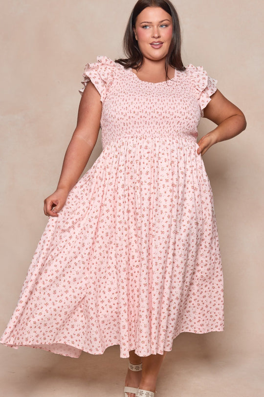 A woman wearing the Hattie Dress in Madeline Pink by Ivy City Co—a light pink, floral-print cotton midi dress with ruffled sleeves—stands against a neutral background, holding out the skirt and smiling.