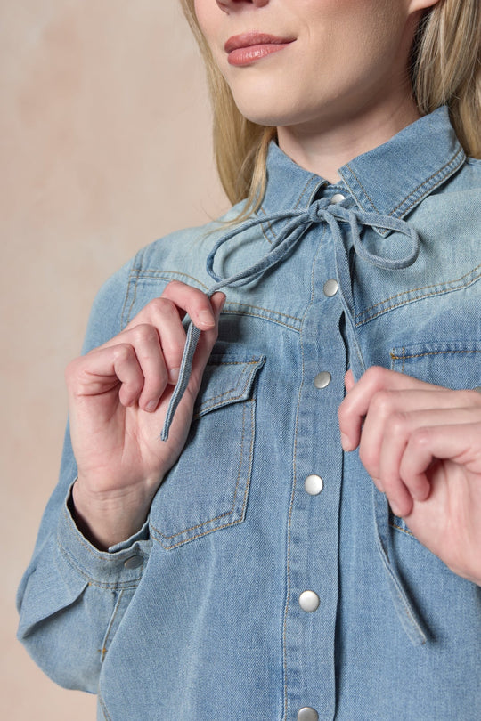 A woman, shown from the lower face to upper torso, adjusts the self-tie bow collar at the neckline of Ivy City Co’s Denver Top in Lightwash Denim—a western-inspired style featuring pearl snap buttons.