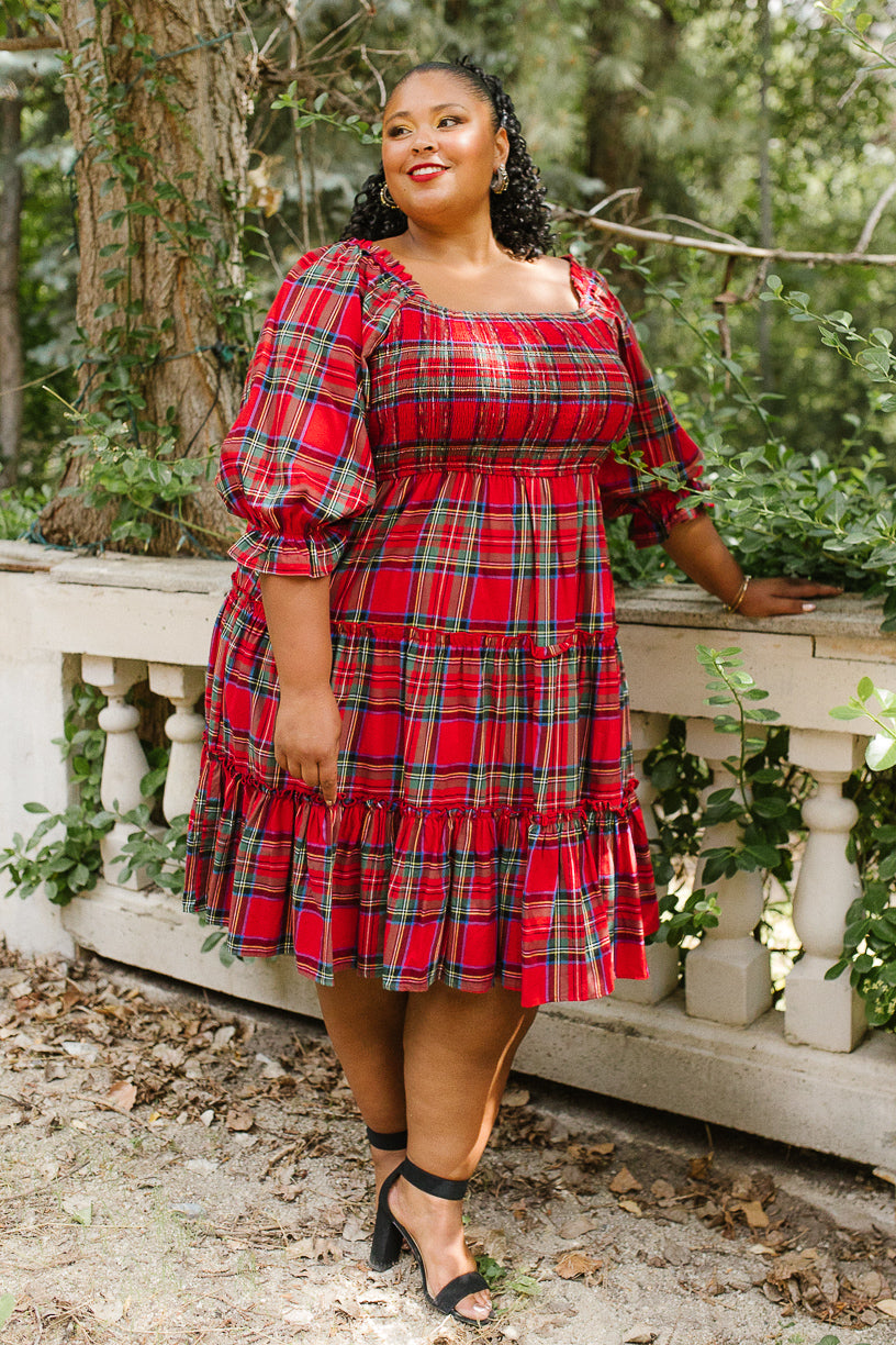 A woman stands outdoors by a white stone railing, surrounded by greenery, wearing the Ivy City Co Madeline Dress in Holiday Plaid. The stylish dress features puffed sleeves and offers a maternity-friendly fit. She is smiling and completes the look with black ankle-strap heels.