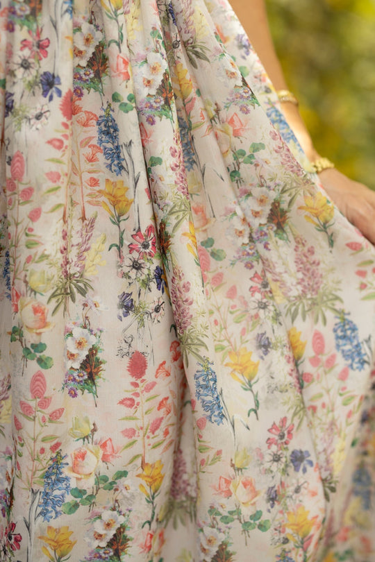 A close-up of the lightweight, flowy fabric from Ivy City Co’s Hattie Dress in Cordelia Floral showcases its vibrant floral pattern, featuring pink, yellow, blue, and green blooms. A hand adorned with gold bracelets gently holds the fabric of this maternity-friendly dress, with a softly blurred background completing the scene.