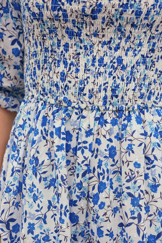 Close-up of the Ivy City Co Madeline Dress in Ditsy Cobalt Floral, showcasing its cotton fabric adorned with various shades of blue flowers and dark stems. The smocked detailing at the bodice adds comfort and elegance, making this midi dress a fresh, versatile choice for any occasion.
