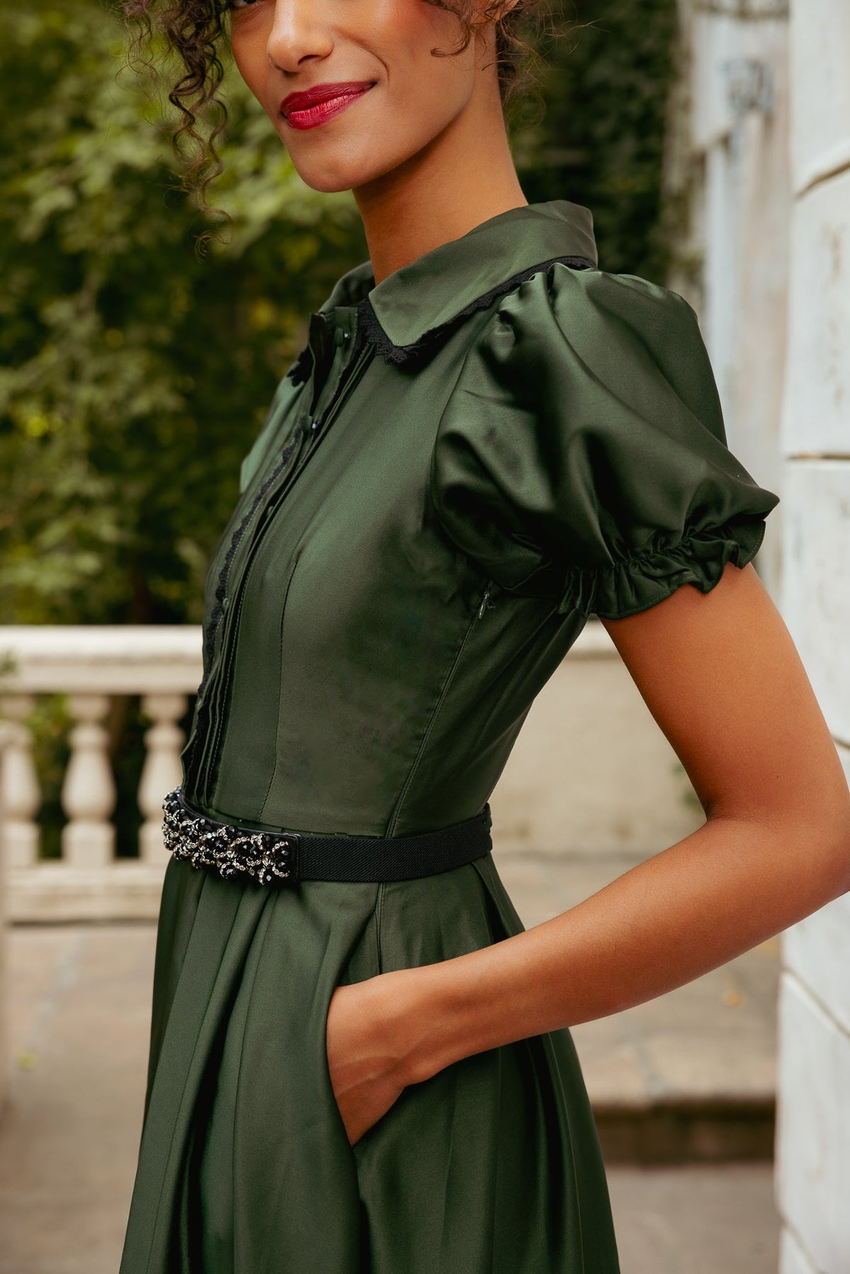 A woman wears a green, short-sleeved Sophia Dress in Green Satin with a black lace-trimmed collar and a black embellished belt.