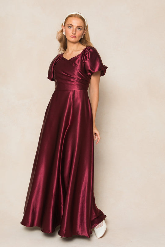 A woman poses confidently against a light beige background, wearing the Tessie Dress in Wine Satin by Ivy City Co. The dress features puff short sleeves and a shimmering satin finish, styled with a white headband and matching shoes—making it the perfect chic choice for fall bridesmaids.