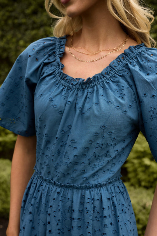 A woman stands outdoors, surrounded by lush greenery, wearing the Ivy City Co Etta Dress in Cobalt—a summer midi dress with eyelet details and short sleeves—styled with two gold chain necklaces.