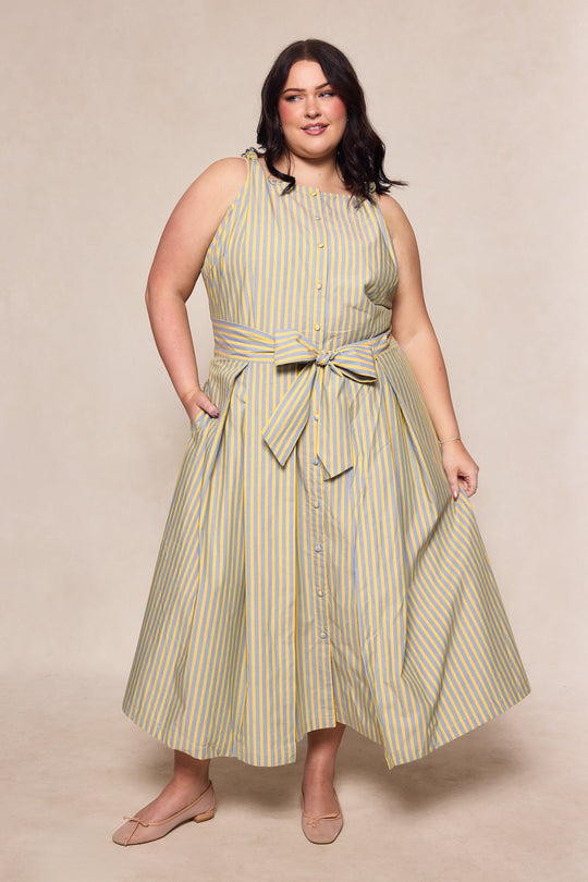 A woman stands smiling with one hand in her pocket, wearing the Glen Dress in Green Stripe by Ivy City Co. This sleeveless, button-down dress features green and white stripes and a waist tie. Designed to be nursing friendly with convenient buttons down the front, it offers both style and practicality. She pairs the look with nude flats and has shoulder-length dark hair.