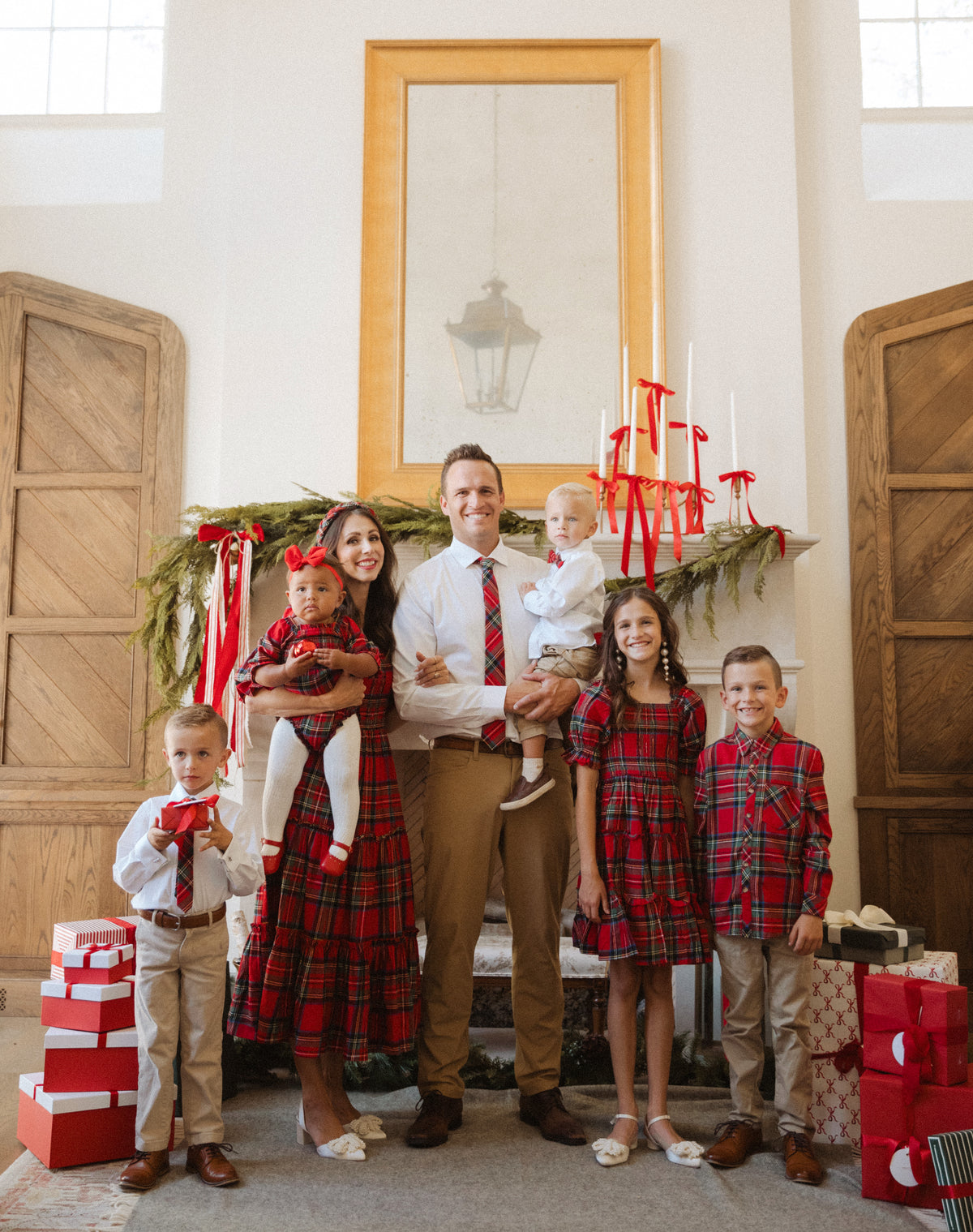 A family of six poses indoors by a decorated fireplace adorned with festive greenery and red ribbons. The children and parents wear coordinated Ivy City Co Madeline Dress in Holiday Plaid outfits, some of which are both maternity friendly and nursing friendly, as they gather around beautifully wrapped presents.