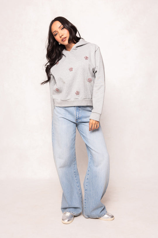 A woman with long dark hair is wearing the Ivy City Co Skylar Hoodie in Heather Gray, featuring small red designs, paired with light blue wide-leg jeans and silver shoes, standing against a plain light background.