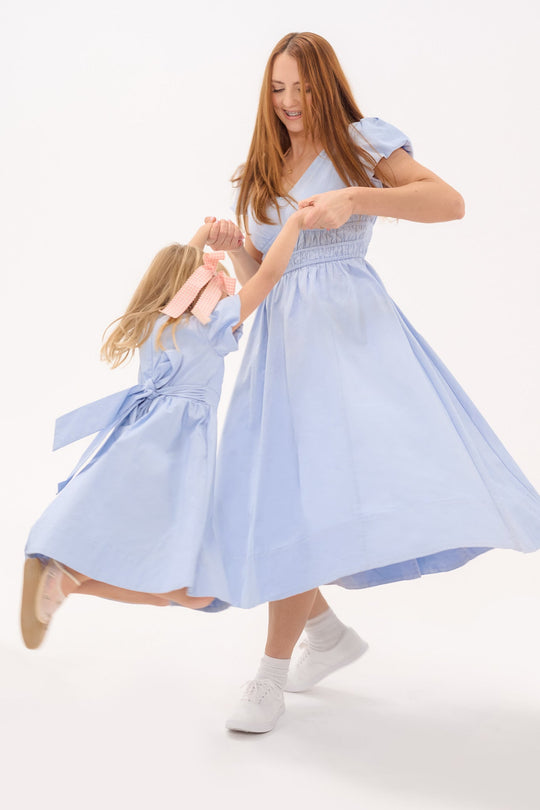A woman and a young girl, both dressed in matching Ivy City Co Zella Dresses in Sky Blue, joyfully dance together holding hands—the girl caught mid-air as the woman smiles—all set against a plain white background.