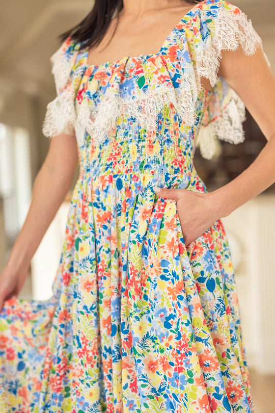 A woman stands indoors in softly lit surroundings, wearing the Ivy City Co Ada Dress in Vibrant Yarrow Print—a colorful floral chiffon dress with white lace trim. She holds part of her skirt and rests one hand in a pocket, drawing attention to the dress’s lively pattern and its nursing-friendly design.