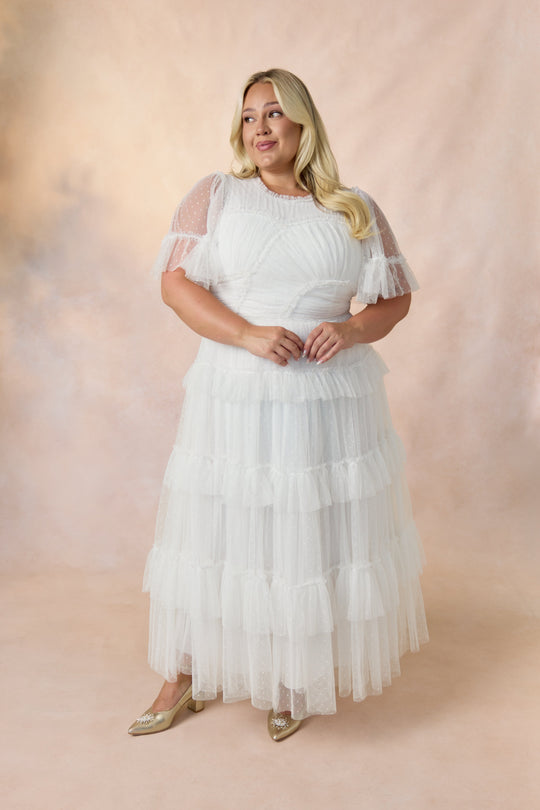 A woman wearing the Whimsical Dress in White by Ivy City Co — a maxi-length gown featuring layered tulle ruffles and sheer sleeves — stands against a soft, light pink and beige backdrop, gently smiling with her hands clasped in front of her.