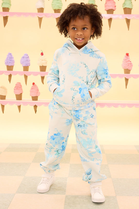 A smiling child stands with hands in pockets, wearing the Ivy City Co Mini Kian Hooded Sweatshirt in Blue Aquarelle Fleur with matching pants. Ice cream cone decorations are displayed in the background.