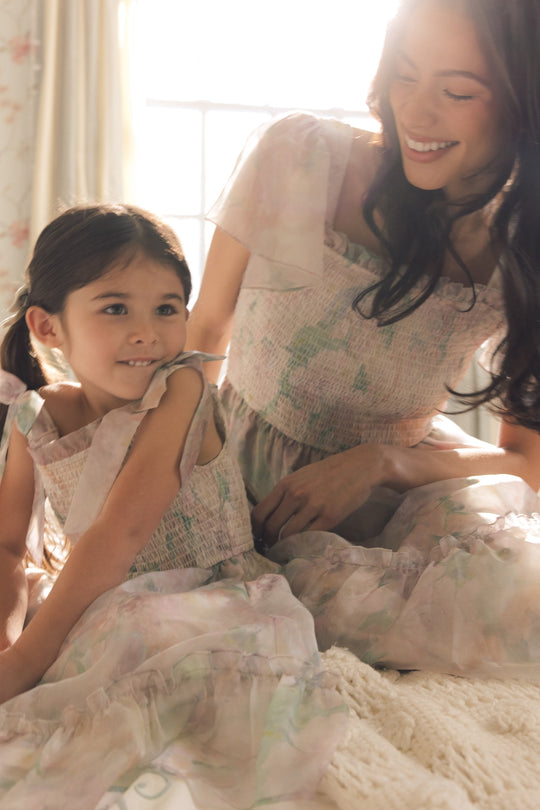 A woman and young girl sit together on a bed, smiling and wearing matching Ivy City Co Rhiannon Dresses in Painted Bloom. Sunlight streams in from a window behind them, creating a warm, soft atmosphere.