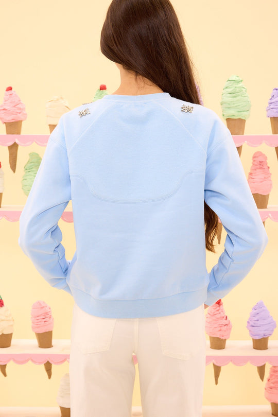 A person with long brown hair, wearing the Ivy City Co Casey Crewneck Sweatshirt in Light Blue with subtle crystal detailing and white pants, stands facing pastel-colored ice cream cone decorations on a cream wall.