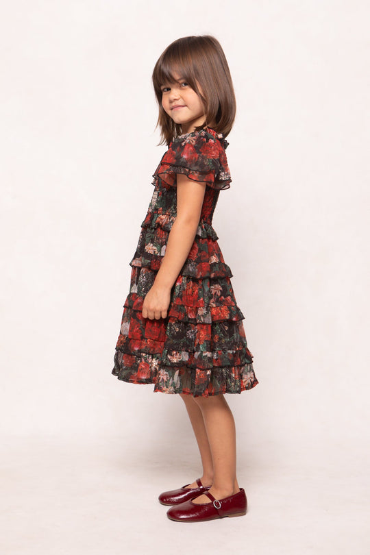 A young girl with straight brown hair stands sideways, smiling. She wears the Ivy City Co Mini Winnie Dress in Midnight Roses—a red and black floral ruffled girls’ maxi dress—paired with matching red shoes, posing against a plain light background.
