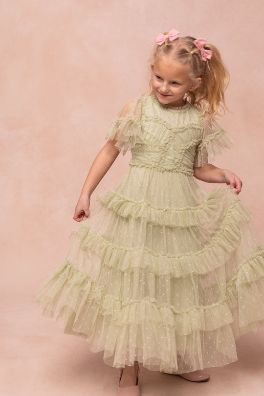 A young girl beams as she holds out her elegant Ivy City Co Mini Whimsical Maxi Dress in Sage, featuring a light green hue with ruffled, layered tulle skirt and sheer sleeves. Pink hair bows adorn her hair as she twirls against a soft beige background.