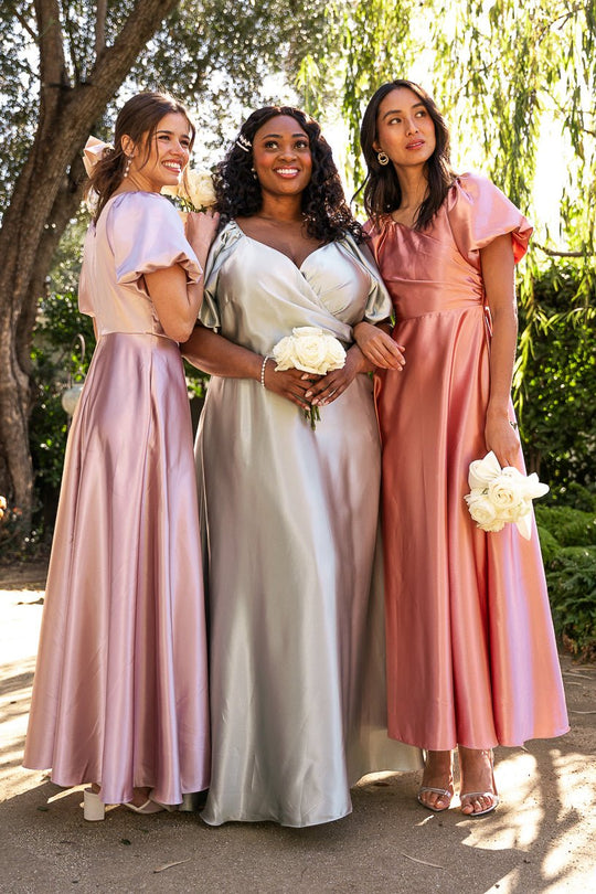 Three women stand outdoors in the sunlight, smiling and holding white roses, each wearing the Tessie Dress in Sage by Ivy City Co. Their modest bridesmaid dresses shimmer in pastel tones as they pose closely together, surrounded by greenery.