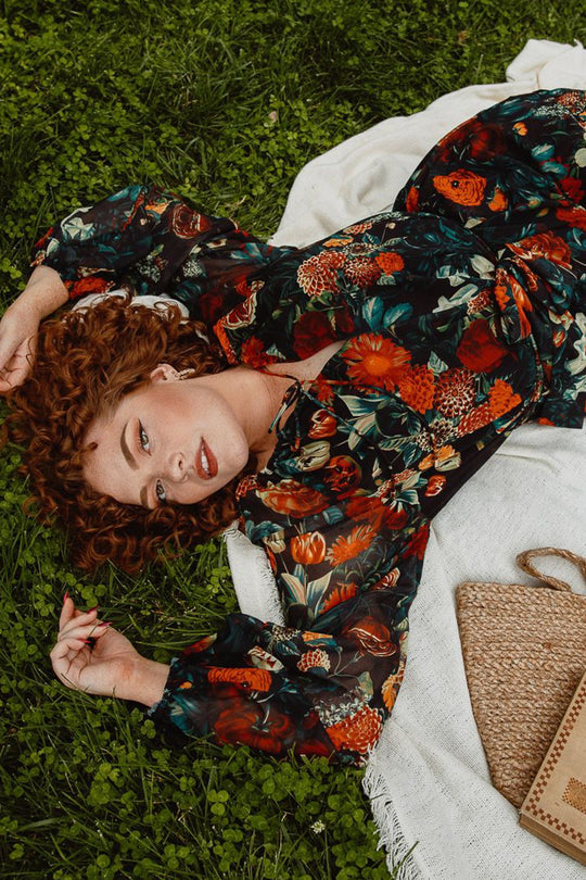 A woman with curly red hair reclines on grass atop a white blanket, wearing the Imogen Dress in Moody Floral by Ivy City Co—a breezy, maternity-friendly floral maxi. She gazes upward with one arm resting above her head, while a woven purse lies nearby.