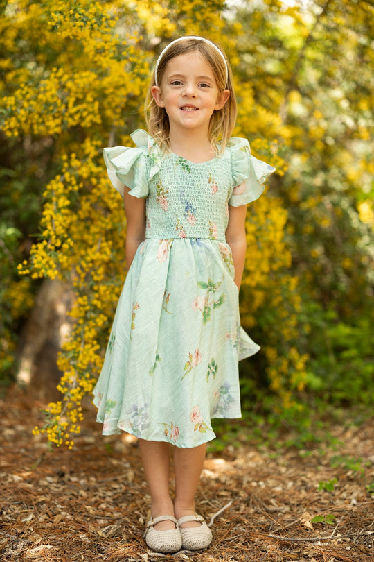 A young girl stands outdoors, smiling in front of yellow flowers and greenery, wearing the Ivy City Co Mini Hattie Dress in Roselyn Blue. Crafted from a soft cotton-linen blend with ruffled sleeves and a smocked bodice, she completes her look with a headband and beige shoes.