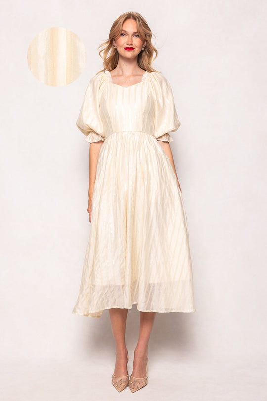 A woman wears the Ivy City Co Tindra Dress in Cream Sequin—a knee-length, puff-sleeve midi with subtle vertical stripes—standing against a plain background and smiling. A small circle in the upper left highlights a close-up of the dress’s fabric texture. This versatile style is maternity friendly and designed to keep you comfortable through every stage.