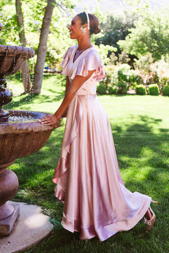 A woman in the Callie Dress in Blush Satin by Ivy City Co stands by a stone fountain in a sunny, green garden, smiling as she touches the fountain’s edge.