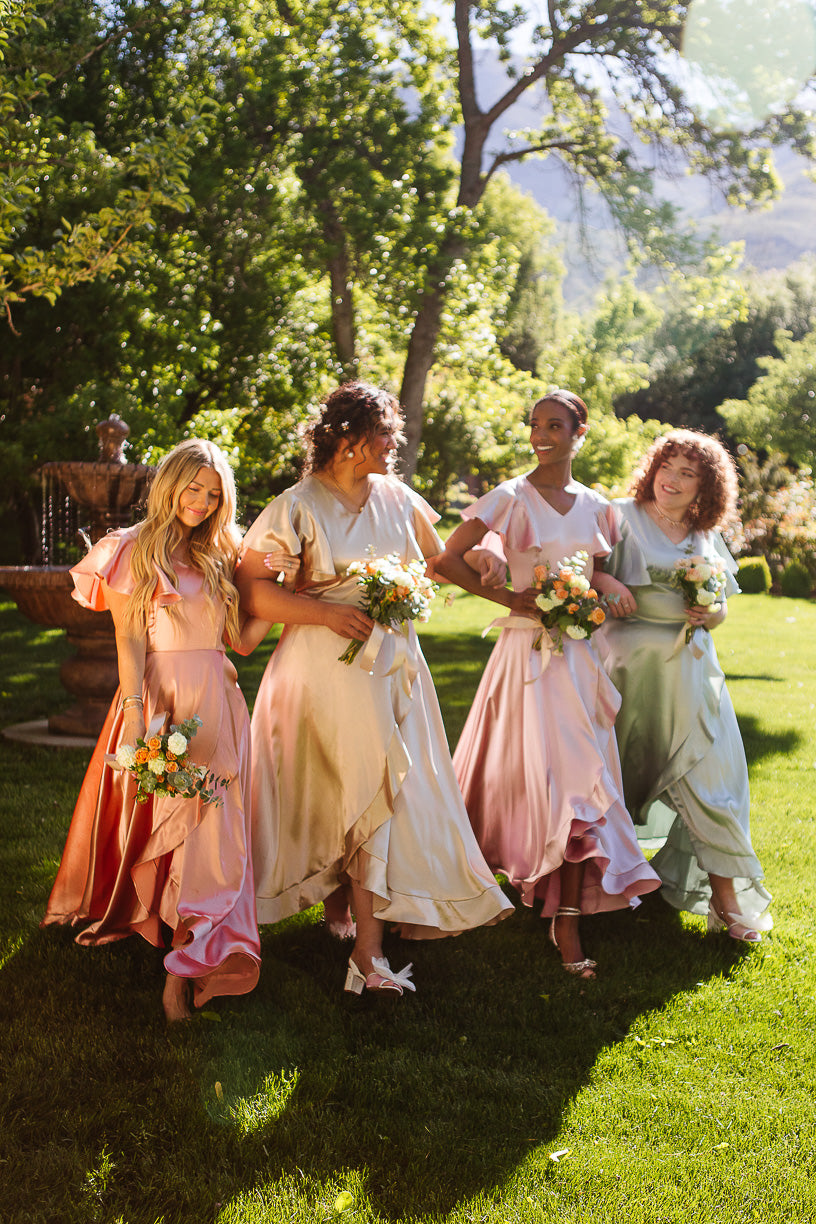 Four women wearing Ivy City Co's Callie Dress in Apricot Crush Satin hold bouquets and walk together on a sunlit lawn, surrounded by greenery, enjoying the outdoor celebration as part of a joyful bridal party.