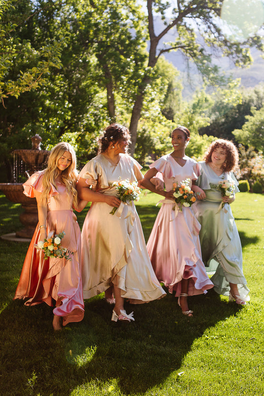 Four women in pastel dresses—one wearing the Ivy City Co Callie Dress in Apricot Crush Satin—hold flower bouquets and smile as they walk together on a sunny, grassy lawn surrounded by trees.