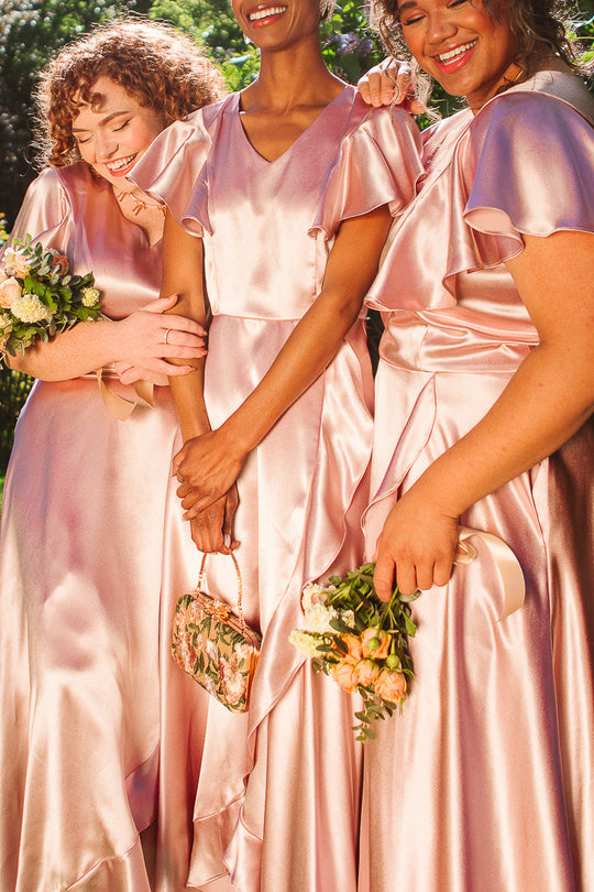 Three women, each wearing the Ivy City Co Callie Dress in Blush Satin, stand close together smiling with small floral bouquets and a floral handbag, surrounded by sunlit greenery.