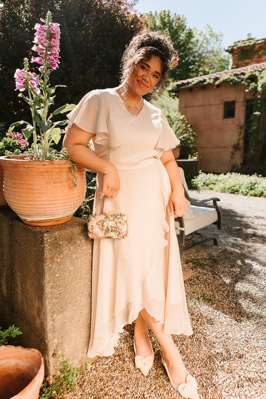 A woman in the Ivy City Co Callie Dress in Champagne Chiffon stands beside a terracotta pot of foxgloves, holding a floral embroidered clutch and wearing bow-detail flats—a beautifully elegant bridesmaid look.