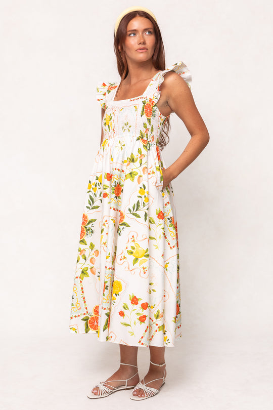 A woman in the Ivy City Co Camilla Dress in Citrus, a white floral sleeveless maxi with a smocked bodice and ruffled straps, stands against a plain background wearing a yellow headband and white strappy sandals.