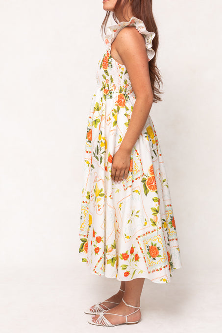 A woman stands sideways against a plain background wearing the Ivy City Co Camilla Dress in Citrus—a white cotton midi dress with a vibrant citrus and floral print, ruffled straps, a smocked bodice, and white strappy sandals.