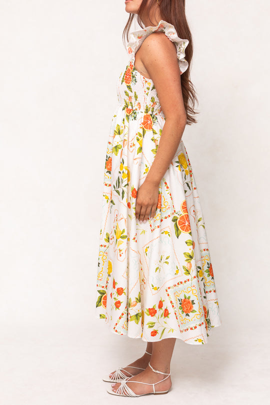 A woman stands sideways against a plain background wearing the Ivy City Co Camilla Dress in Citrus—a white cotton midi dress with a vibrant citrus and floral print, ruffled straps, a smocked bodice, and white strappy sandals.