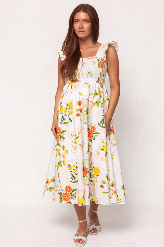 A woman with long brown hair wears the Ivy City Co Camilla Dress in Citrus, a cotton midi dress with a vibrant citrus print and ruffled straps, paired with white strappy sandals against a plain light background.