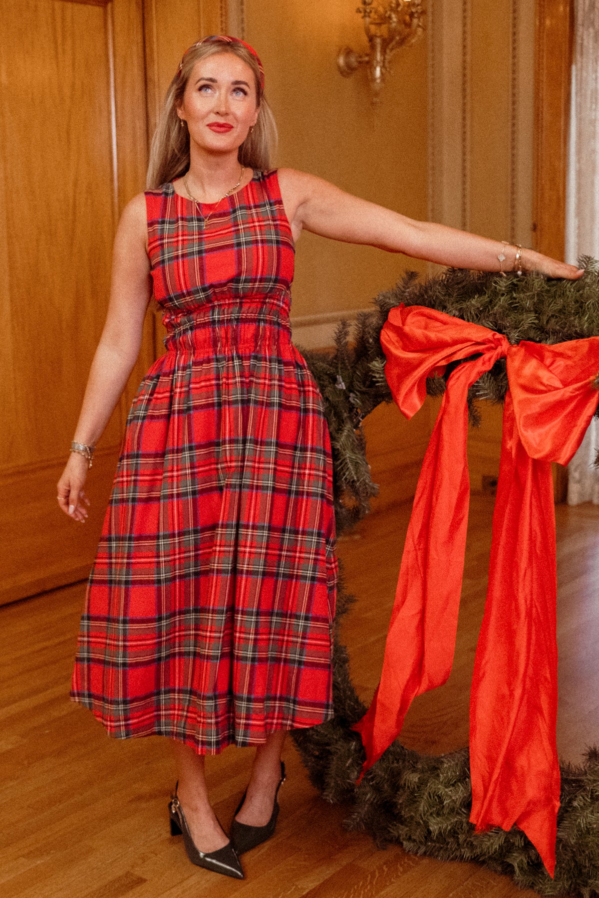 A woman with long blonde hair, wearing a red headband and black heels, stands indoors on a wooden floor holding a large holiday wreath adorned with a red bow. Her stylish and maternity-friendly Carly Dress in Holiday Plaid by Ivy City Co completes her festive look.