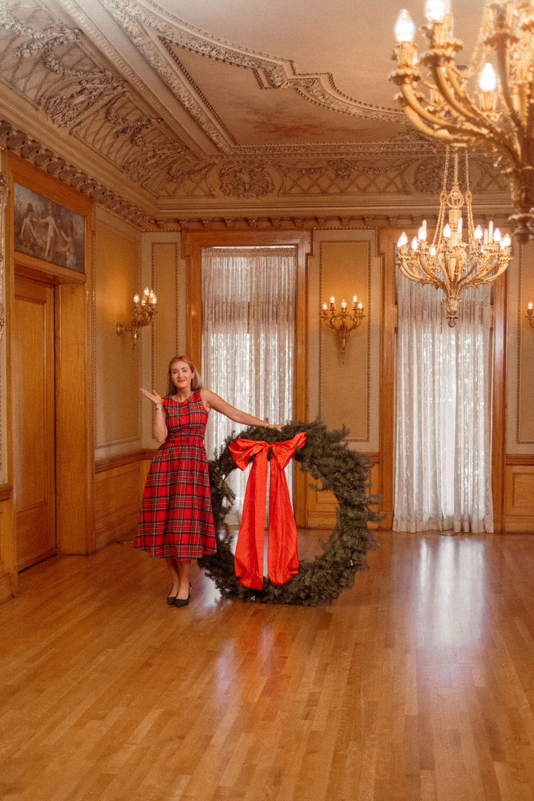 A woman wearing the Carly Dress in Holiday Plaid by Ivy City Co stands in an elegant room, holding a giant holiday wreath adorned with a large red bow. The chandeliers and ornate moldings provide a festive backdrop for this maternity-friendly Christmas dress.
