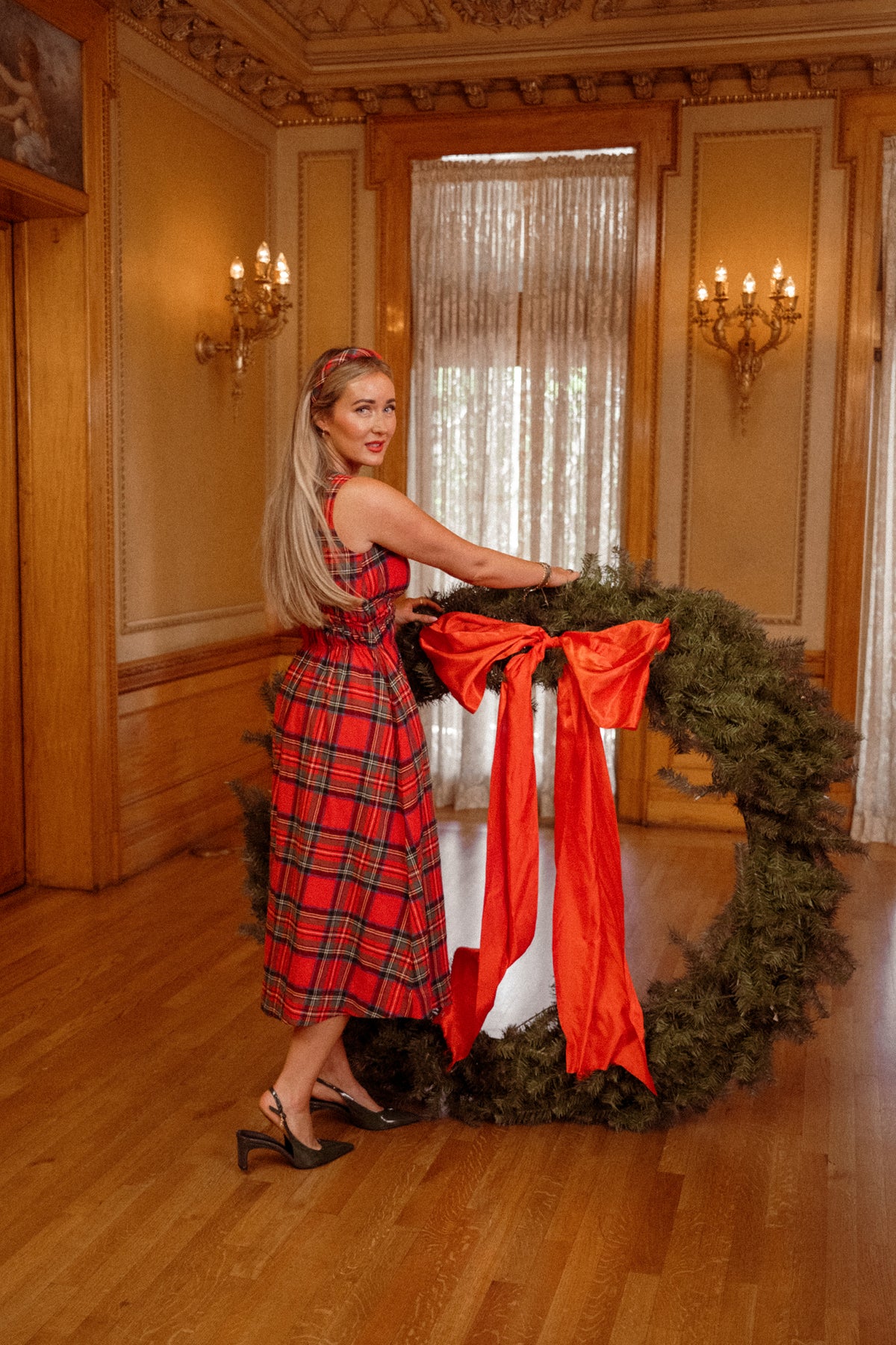 A woman wearing the Ivy City Co “Carly Dress in Holiday Plaid” stands indoors, holding a large green wreath adorned with a big red bow. The elegant room features wooden floors, ornate walls, and tall windows draped with sheer curtains.