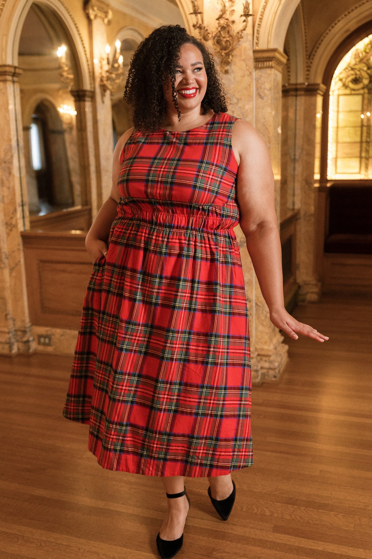 Wearing the Carly Dress in Holiday Plaid by Ivy City Co, a woman in a sleeveless red plaid dress and black heels stands in an elegant room with ornate columns, wooden floors, and arched windows, smiling and playfully holding one hand out.