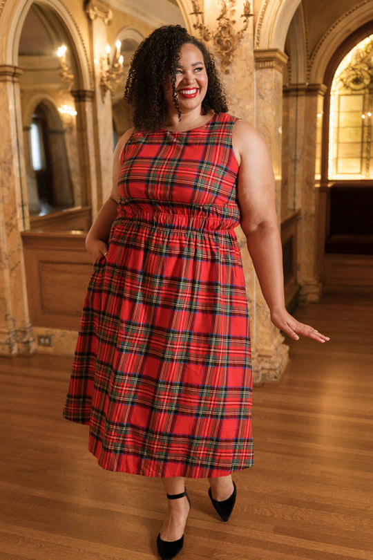 Wearing the Carly Dress in Holiday Plaid by Ivy City Co, a woman in a sleeveless red plaid dress and black heels stands in an elegant room with ornate columns, wooden floors, and arched windows, smiling and playfully holding one hand out.