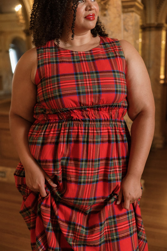A woman stands indoors, gently holding the fabric of her Ivy City Co Carly Dress in Holiday Plaid. Only the lower half of her face and body are visible, drawing attention to the chic, maternity-friendly design.