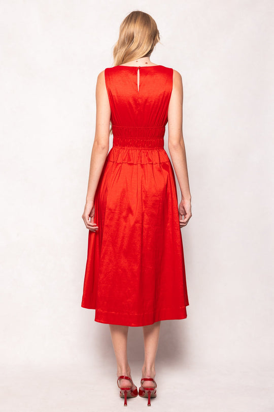 A woman with long blonde hair stands facing away against a light background, wearing the sleeveless, knee-length Carly Dress in Red Taffeta by Ivy City Co, styled with red high-heeled sandals and featuring a fitted waist.