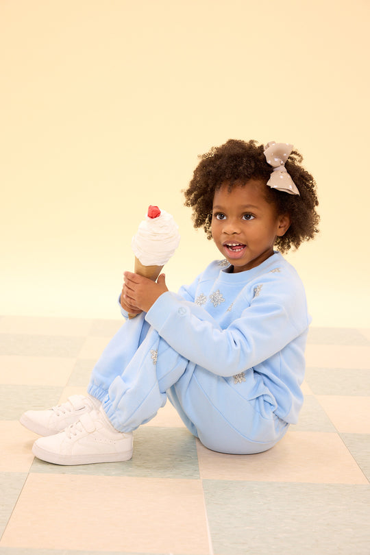 Child in a light blue outfit holding a toy ice cream cone on a checkered floor.
