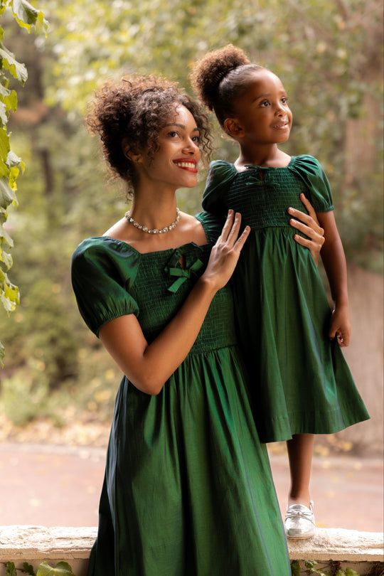 Woman and young girl in matching green dresses standing outdoors with greenery in the background