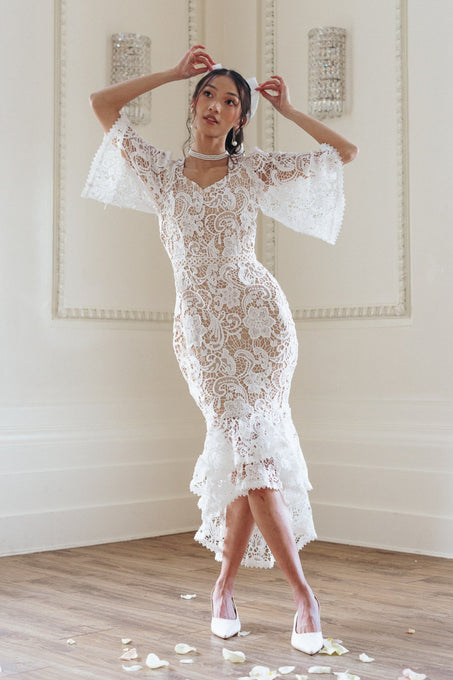 A woman poses indoors wearing the Cecilia Dress in White Lace by Ivy City Co, featuring flared sleeves and a ruffled hem. She pairs it with white heels and a choker, while rose petals are scattered on the wooden floor beneath her.