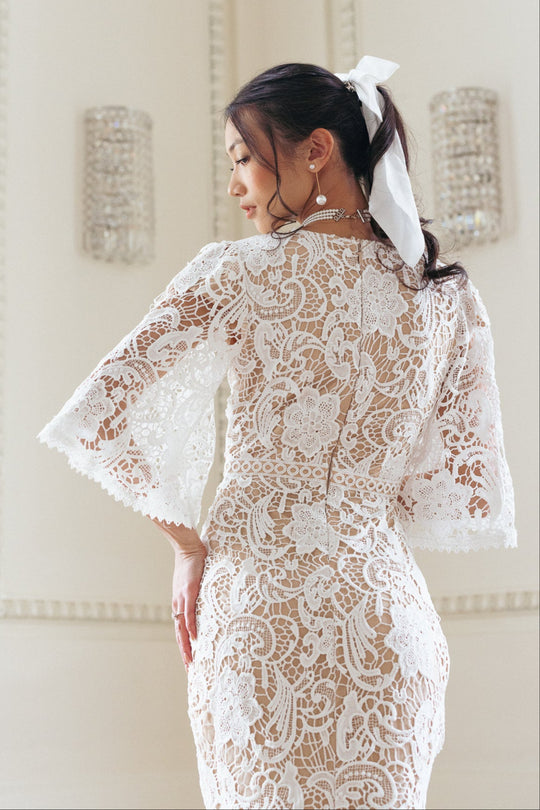 A woman, her dark hair tied with a white ribbon, stands with her back to the camera wearing the Ivy City Co Cecilia Dress in White Lace—featuring pockets—in a softly lit room with ornate walls and crystal sconces.