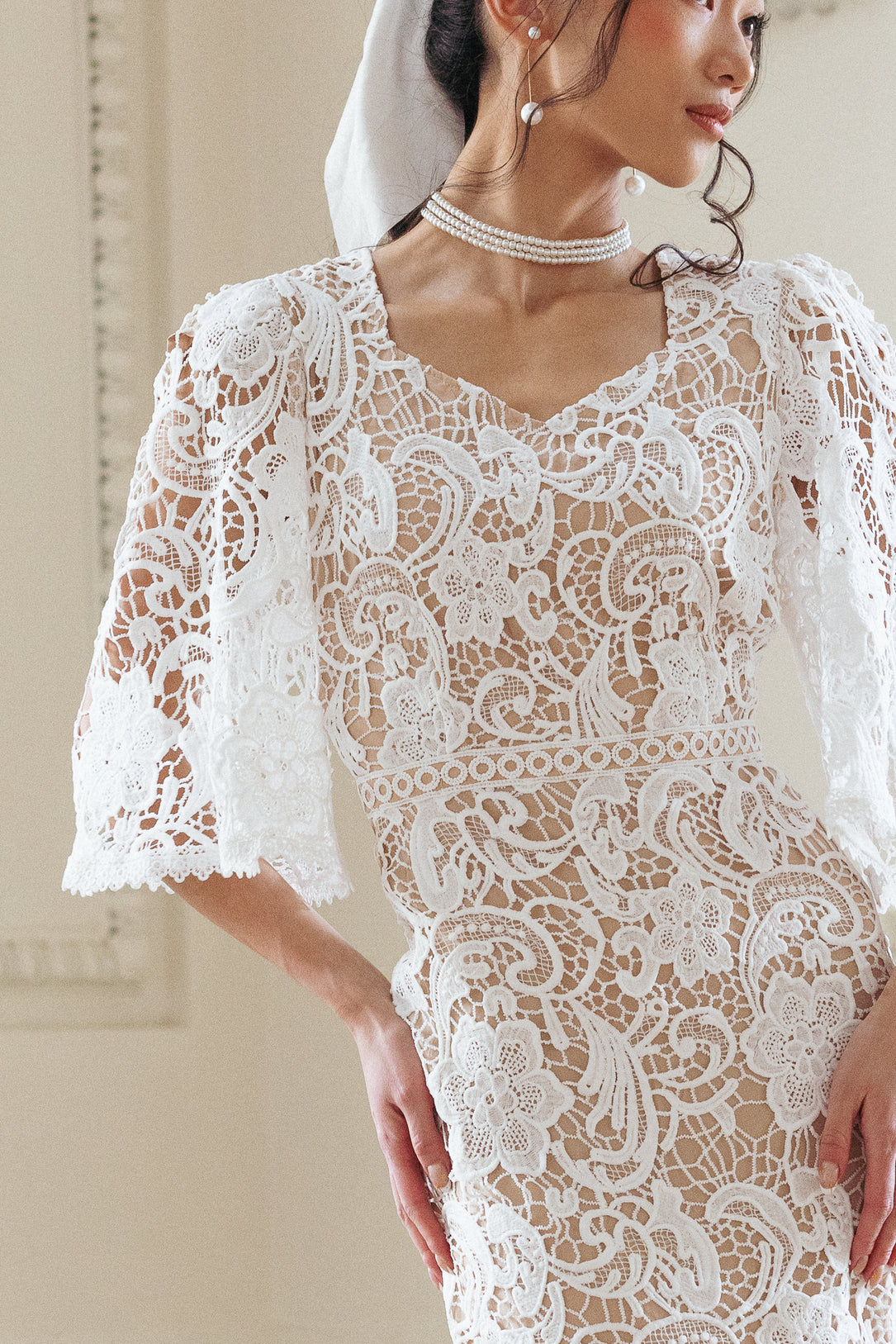 A woman wears the Ivy City Co Cecilia Dress in White Lace, featuring floral lace, puffed sleeves, and a midi silhouette. She accessorizes with pearl earrings, a pearl necklace, and a veil over her updo. Only her upper body and part of her face are visible.