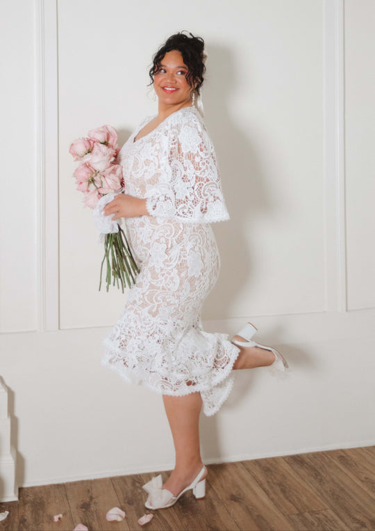 A woman poses playfully in the Cecilia Dress in White Lace by Ivy City Co, smiling with a raised leg and holding pink roses in a bright room with light walls and wooden floors.