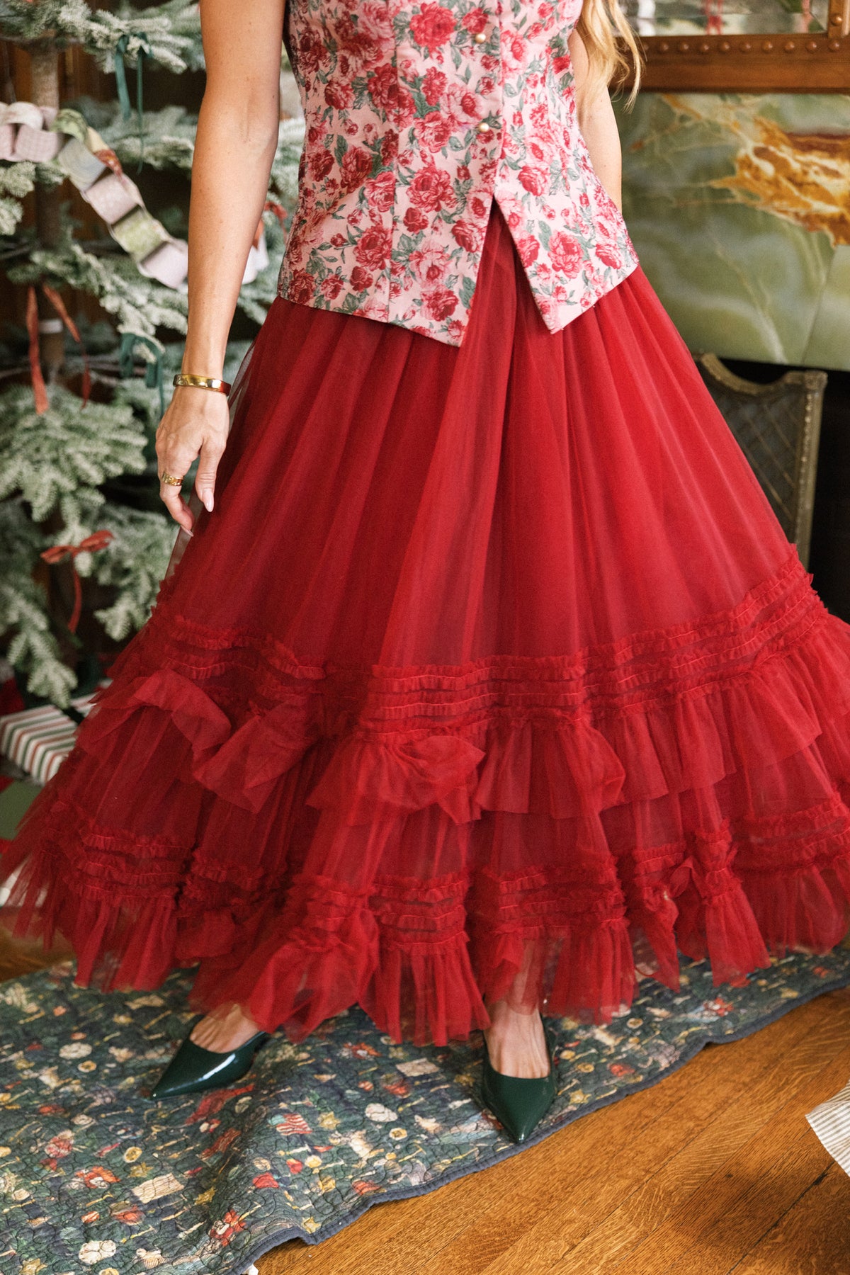 Wearing the Ivy City Co Celestine Skirt in Garnet, a person stands on a colorful rug beside a decorated tree and wrapped gifts, with only the lower half visible.