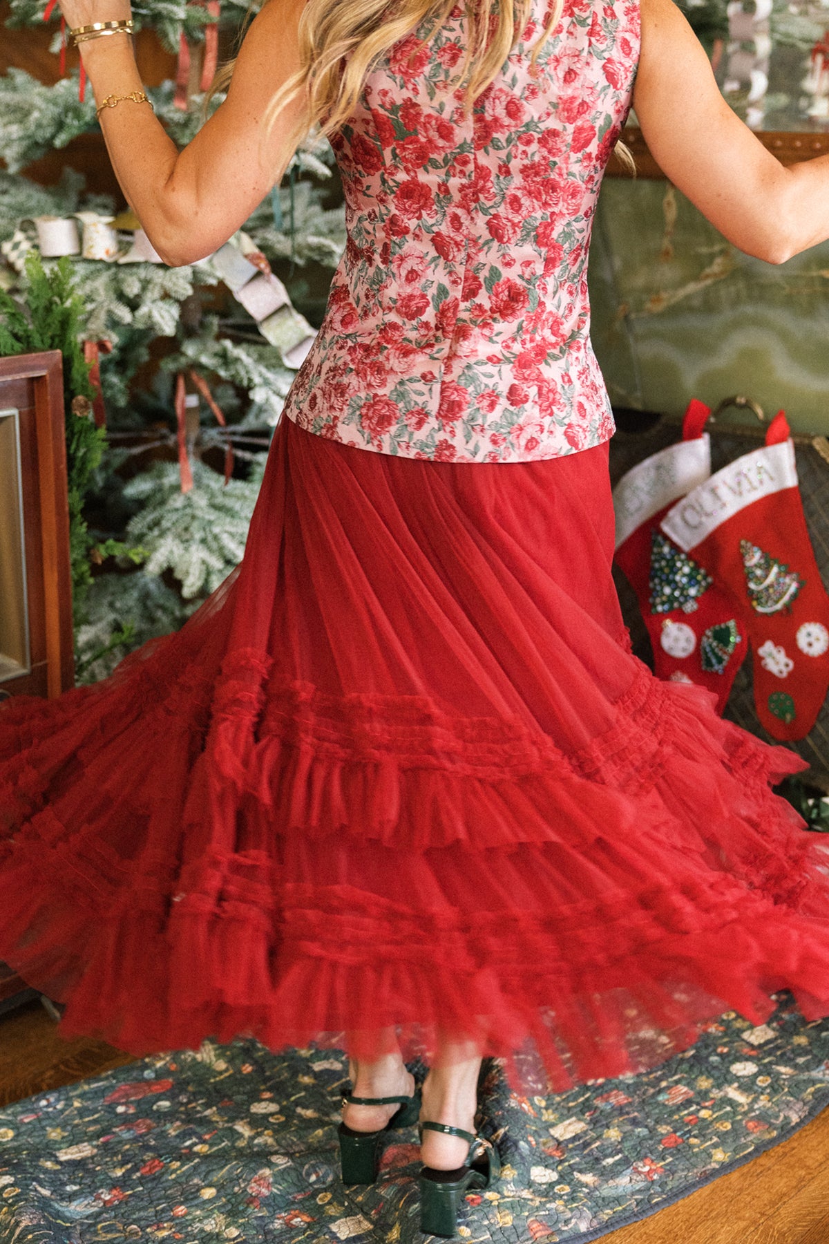 A woman, seen from behind, twirls indoors near a decorated Christmas stocking and tree, wearing the Ivy City Co Celestine Skirt in Garnet and a floral top.