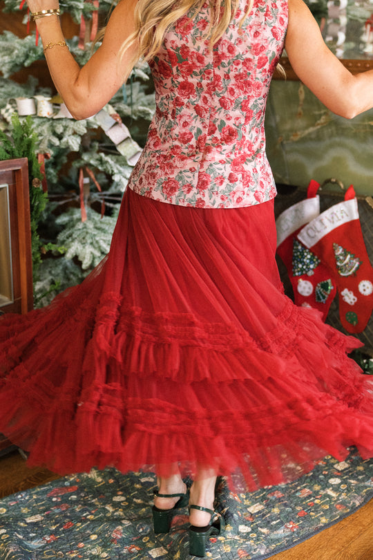 A woman, seen from behind, twirls indoors near a decorated Christmas stocking and tree, wearing the Ivy City Co Celestine Skirt in Garnet and a floral top.