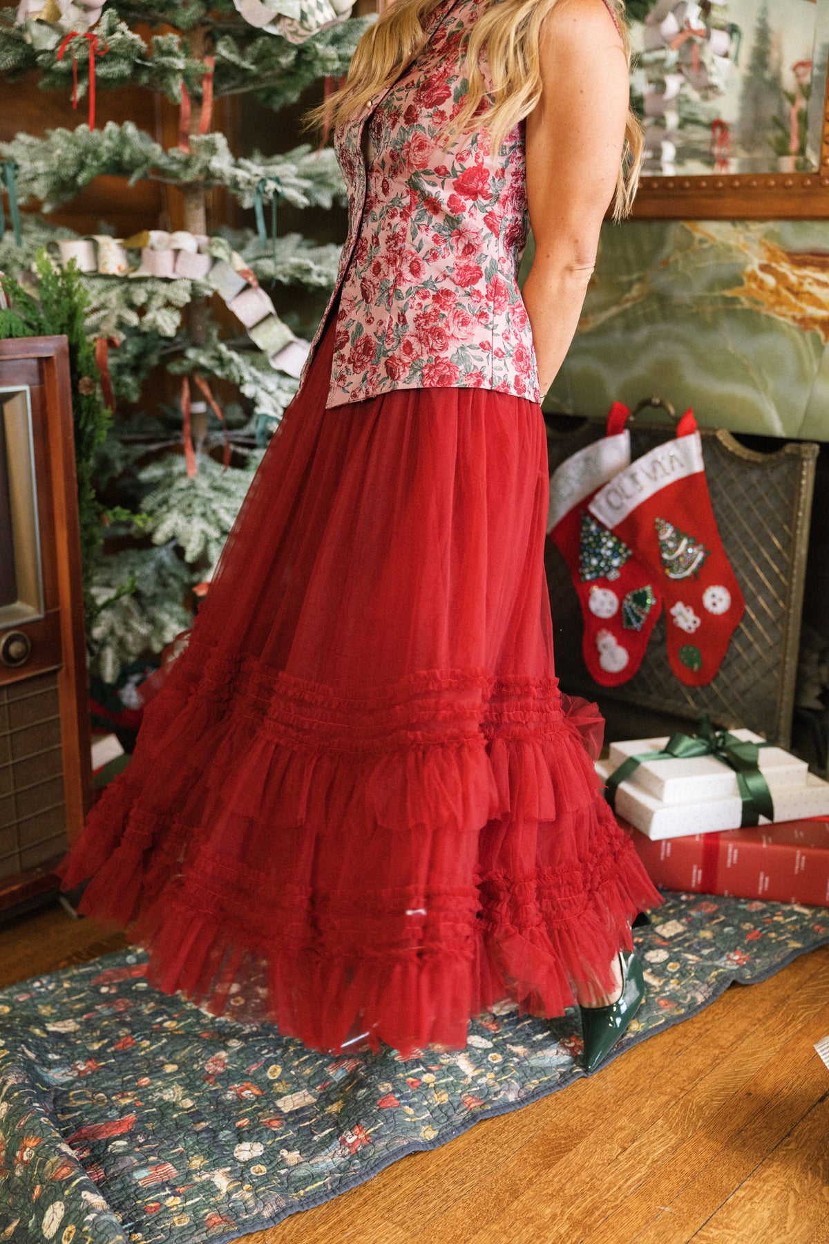 A woman in a floral top and the Ivy City Co Celestine Skirt in Garnet stands by a Christmas tree, surrounded by stockings, wrapped gifts, and a vintage radio. Her face is not visible.