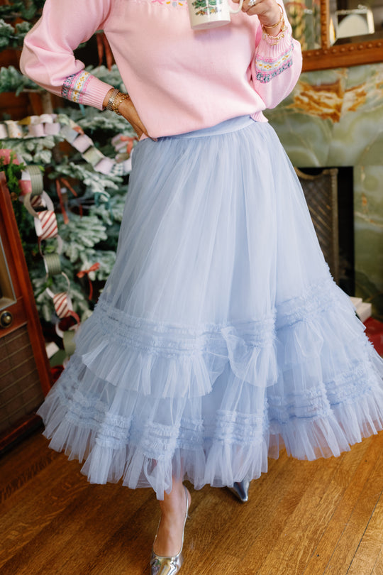 Wearing Ivy City Co’s Celestine Skirt in Misty Blue paired with a cozy pink sweater, a person stands indoors on a wooden floor, holding a mug. Only the outfit and lower face are visible, creating the perfect look for a holiday recital.